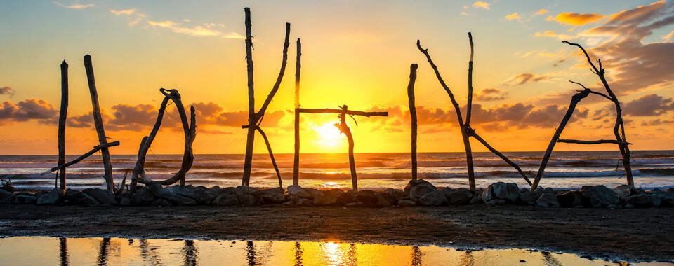 Driftwood poles arranged to spell 'HOKITIKA' stand along a rocky barrier at the beach during sunset, with a warm orange sky and the ocean in the background and their reflection visible in wet sand.