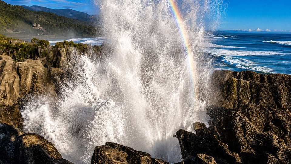 Ocean waves crash against jagged rocks with a burst of spray forming a rainbow overhead and blue sky beyond the coastgrouped scene