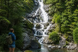 Hiker with backpack standing at a rocky pool, watching a multi-tiered waterfall in a green forest.