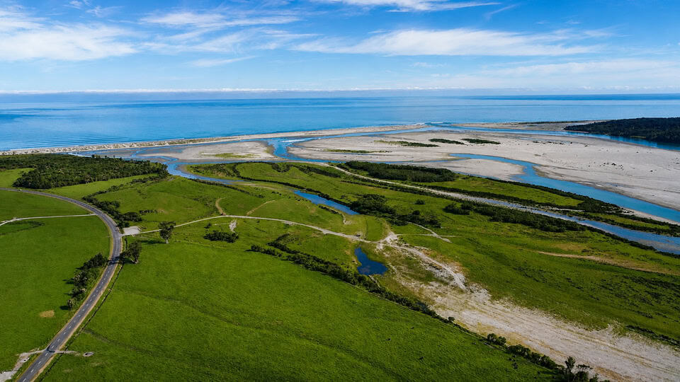 Aerial view of a coastal landscape: green fields with winding roads and small streams leading to a sandy shoreline and calm blue ocean under a clear sky.