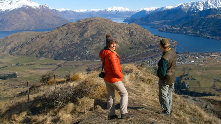 looking-over-queenstown-and-lake-wakatipu-29 | Must Do New Zealand | Must Do New Zealand Two hikers stand on a grassy hill overlooking a blue lake and snow-capped mountains in the distance.