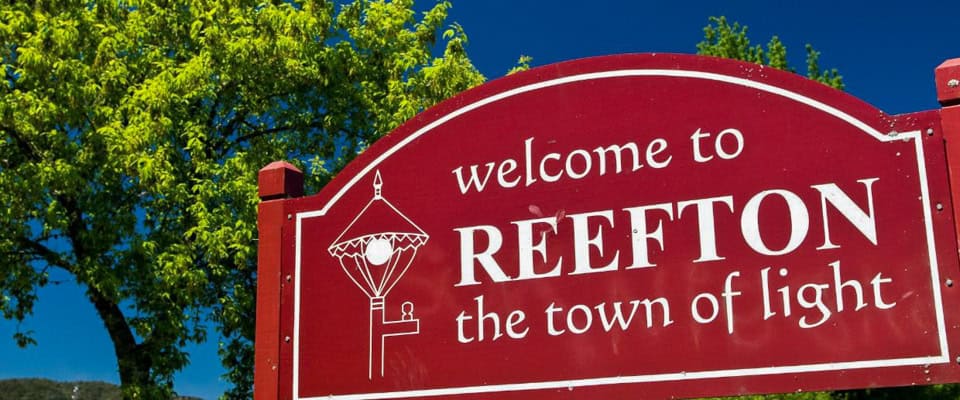 Red welcome sign for Reefton reading 'welcome to REEFTON the town of light' against a blue sky with green trees nearby (informational)