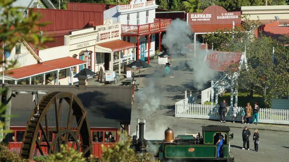 Green steam locomotive on a historic western town street with wooden storefronts, a water wheel, and bystanders.