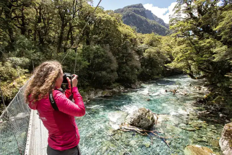 Woman in a pink jacket takes a photo from a railing bridge over a clear river in a forested area.