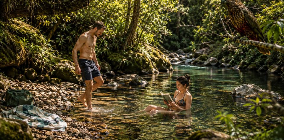 Man in swim shorts stands in a shallow tropical river as a woman sits in the water reading a book, with dense greenery and a parrot perched above.