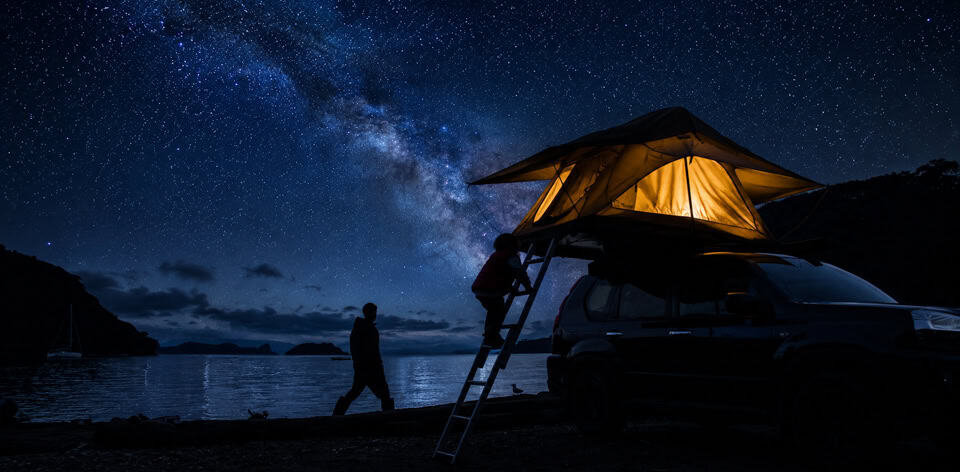 Night camping by the water with a glowing rooftop tent atop a car under a starry night sky and Milky Way.