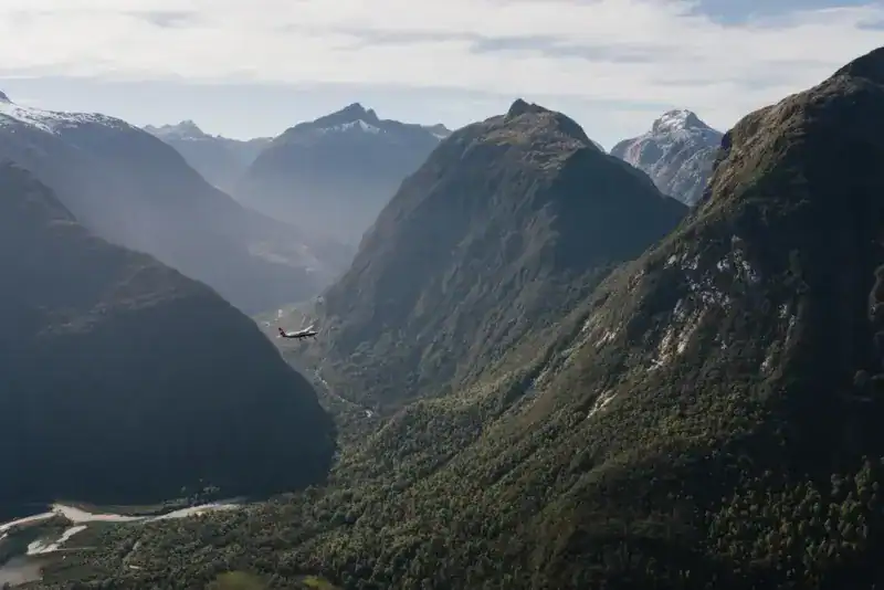 Small red airplane flying through a deep green valley between towering mountains, with a river visible at the valley floor.