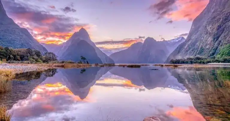 Tranquil alpine lake at sunset, jagged mountains surround the calm water, pink/orange sky reflected in the surface.