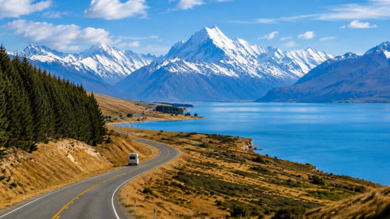 queenstown-to-Christchurch Drive-33 | Must Do New Zealand | Must Do New Zealand Winding coastal road beside a blue lake with snow-capped mountains in the distance and evergreen trees on the left.